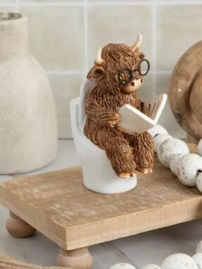Whimsical Brown Highland Cow Figurine Reading on White Toilet
